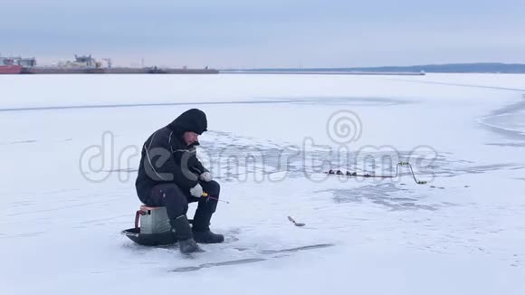 穿着深色衣服的老渔夫在河港的背景下在冰冻的河面上用冬季的鱼竿钓鱼视频的预览图