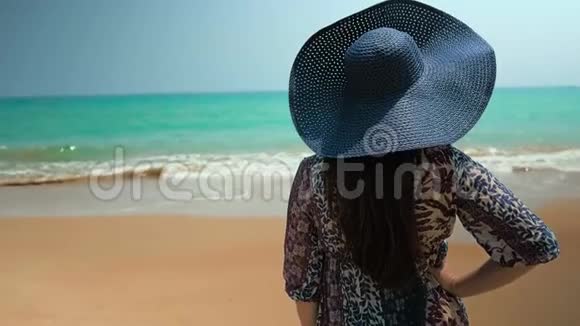 海滩上的女孩戴着蓝色的帽子和蓝色的裙子看着海洋近距离射击慢动作视频的预览图