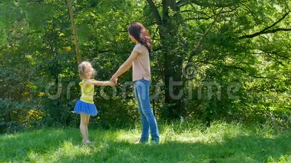在公园里穿着粉色衬衫的年轻母亲和穿着牛仔裤旋转可爱的小女儿的户外画像视频的预览图