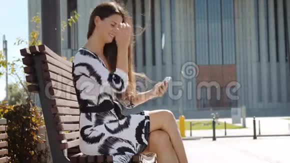 年轻的商务女性穿着连衣裙手里拿着电话坐在长凳上工作视频的预览图