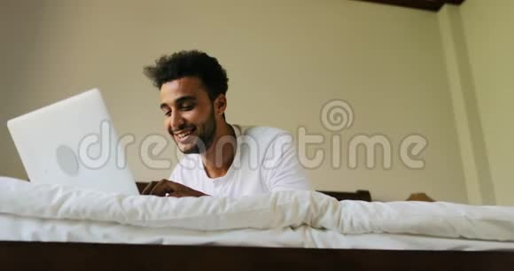 年轻人用笔记本电脑躺在床上快乐地微笑着西班牙男人在床上聊天视频的预览图