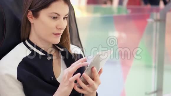 年轻女性使用手机浏览在购物中心舒适的皮椅上放松视频的预览图