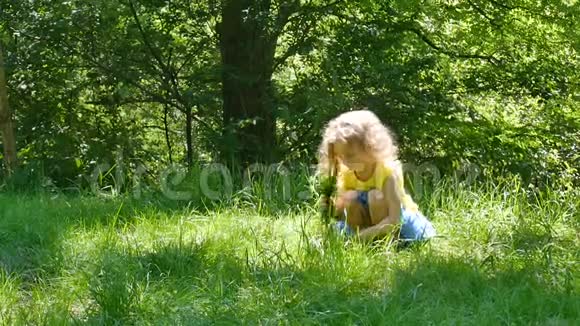 小可爱的金发女孩穿着明亮的黄色衬衫和蓝色的裙子坐在公园里的绿草上把它捡起来视频的预览图