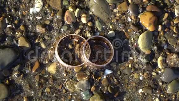 两个金色的结婚戒指躺在沙滩上的石头上阳光照耀着接近宏观环上的光的灌注海洋视频的预览图