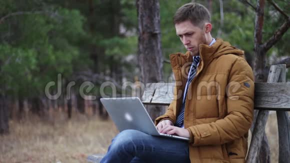 那个穿着领带和夹克的人正坐在一个秋天公园的长凳上拿着一台笔记本电脑在键盘上输入文字冲浪视频的预览图