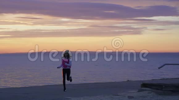 下面的动作年轻苗条的女孩戴着运动帽和运动衫早上在大河堤上慢跑视频的预览图
