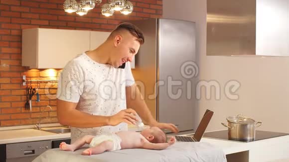 年轻的父亲在厨房喂婴儿在笔记本电脑上工作视频的预览图