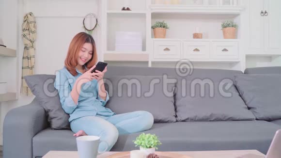 美丽迷人微笑的亚洲女人的肖像她用智能手机拿着一杯温暖的咖啡或茶躺在沙发上视频的预览图