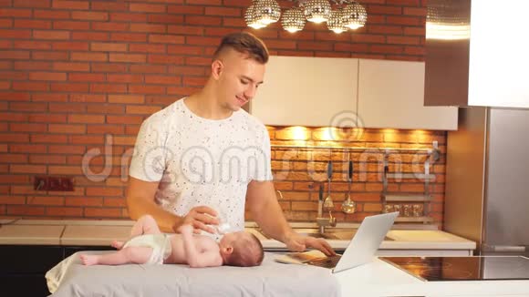 年轻的父亲在厨房喂婴儿在笔记本电脑上工作视频的预览图