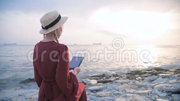 日落时分一个穿着夏红色连衣裙和草帽坐在海边的一块石头上的迷人少女视频的预览图