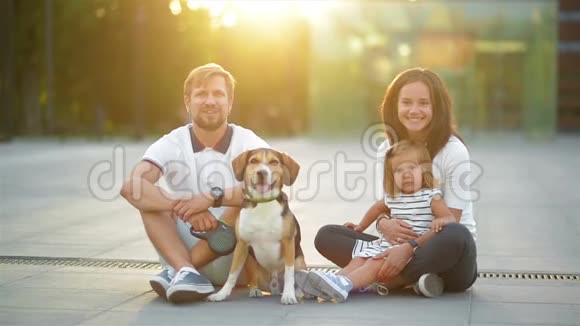 夏季日落时分在城市里带着小女儿和他们的小猎犬的年轻家庭的户外肖像视频的预览图