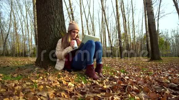 迷人的年轻女孩在秋天公园的树下坐在地上在笔记本电脑里喝咖啡视频的预览图