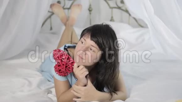 漂亮的年轻女子穿着蓝色连衣裙在床上玩得很开心拿着一个心形玩具视频的预览图