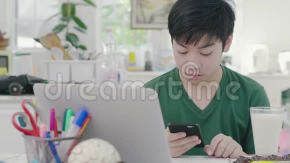 亚洲小男孩在智能手机上使用卷轴同时慢慢地吃有机牛奶早餐视频的预览图