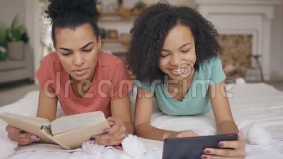 快乐的混血有趣的女孩使用平板电脑向她的妹妹展示平板电脑上的照片她在看书视频的预览图