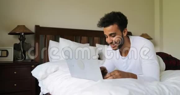 年轻人用笔记本电脑躺在床上快乐地微笑着西班牙男人在床上聊天视频的预览图