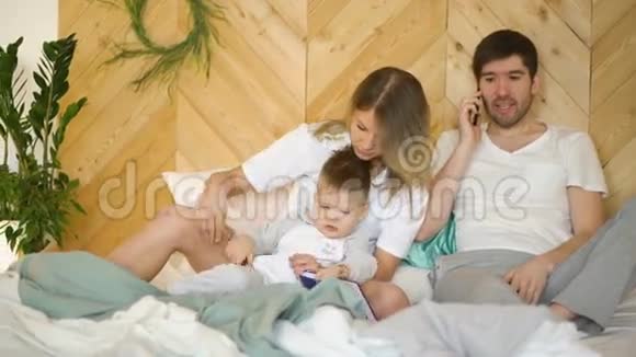 年轻的母亲和儿子玩而父亲在床上打电话视频的预览图