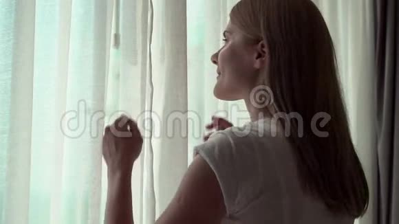 穿白色t恤的女人揭开窗帘看着窗外欣赏100英尺的海景视频的预览图