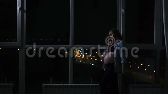孕妇在晚上用手机说话摸着他的肚子站在大的全景窗口视频的预览图