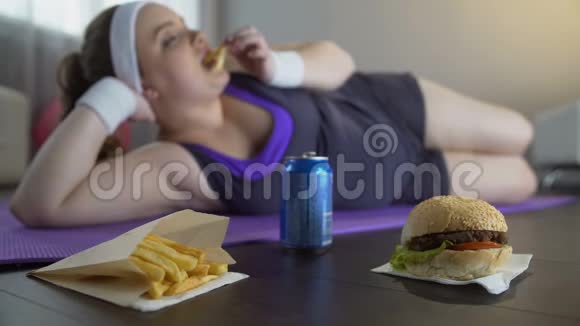 懒女人穿着运动服躺在瑜伽垫上吃垃圾食品而不是训练视频的预览图