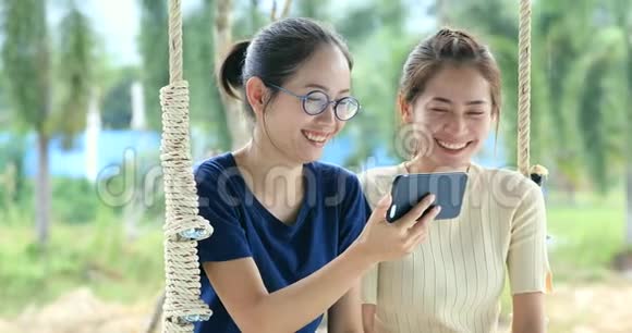 快乐的亚洲家庭大姐和妹妹手拿手机在秋千椅上微笑着面对视频的预览图