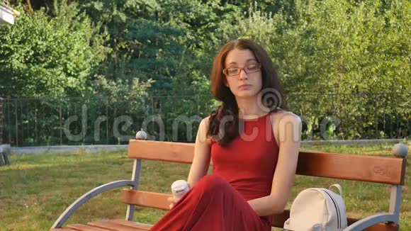 穿着深红长衣和眼镜的可爱的女青少年喝着坐在长凳上的热杯视频的预览图