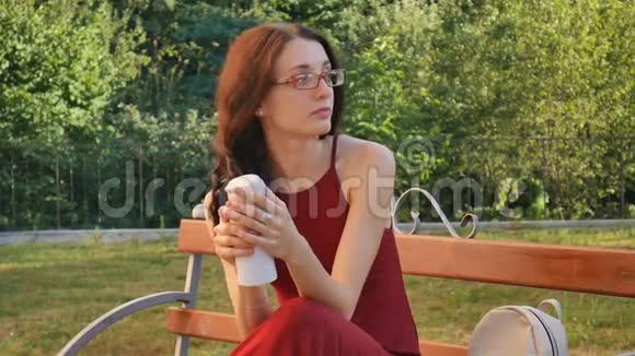 戴着眼镜的美丽的深色头发女孩坐在长凳上的户外带着饮料瓶和饮料视频的预览图