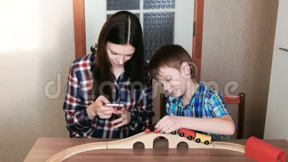 一起玩妈妈看着电话儿子正在玩一条木制铁路火车马车和隧道坐在那里视频的预览图