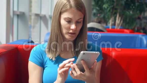 一幅美丽梦幻的年轻女子穿着蓝色t恤坐在咖啡馆里思考的肖像她的智能手机视频的预览图