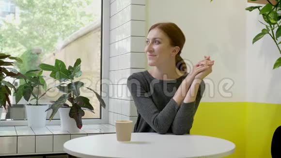 年轻的美女扎着马尾辫坐在咖啡馆里看着窗外和镜头微笑着视频的预览图