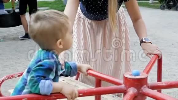 在公园的彩色旋转木马上年轻母亲骑着她蹒跚学步的孩子的慢动作镜头视频的预览图