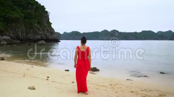 那个穿红色衣服的女人在海滩上散步那个穿红色衣服的女人走在岛上的海岸上后视图视频的预览图