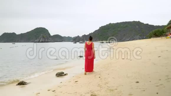 那个穿红色衣服的女人走在岛上的海岸上后景海洋冲浪热带岛屿沙子上的腿迹视频的预览图