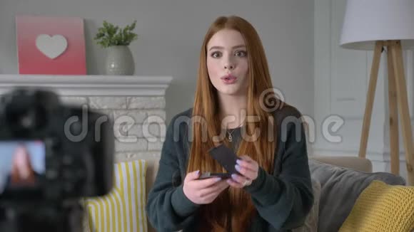 年轻的红发女孩博客作者微笑着对着镜头说话展示了一个新的购买化妆品眼影托盘家视频的预览图