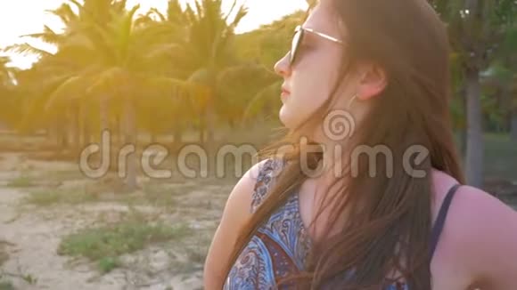 戴着蓝色帽子和眼镜的年轻女孩看着远处在热带棕榈树的背景下微笑慢动作视频的预览图