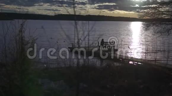 日落时分一个女孩坐在湖边的木甲板上的长椅上视频的预览图