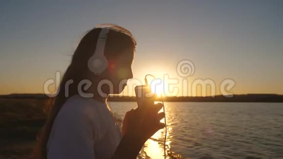 女孩戴着耳机在日落时带着手机靠近水面慢动作视频的预览图