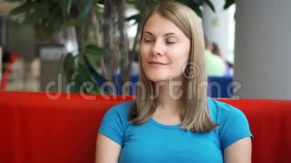 一幅美丽梦幻的年轻女子穿着蓝色t恤坐在咖啡馆里思考的肖像红沙发青树视频的预览图