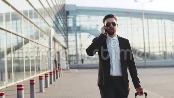 年轻的时尚男士戴着太阳镜在手机上说话离开机场时拉着手提箱成功的生活视频的预览图
