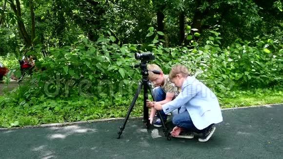 两个男孩把摄像机放在绿色公园背景的三脚架上视频的预览图