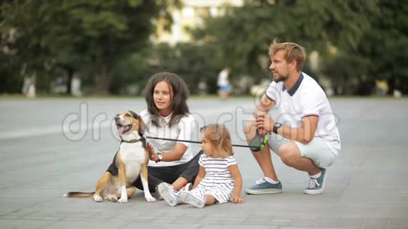 漂亮的年轻母亲留着胡子的父亲可爱的小女儿和小猎犬正坐在城市广场上视频的预览图