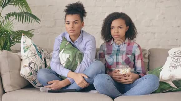 两个混血卷发女孩朋友坐在沙发上看紧张的电视节目在家吃爆米花视频的预览图