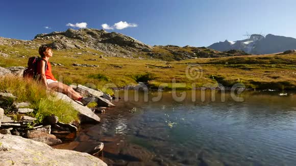红头发的女人带着背包在高山湖畔享受轻松的日光浴阿尔卑斯山壮丽的全景视频的预览图
