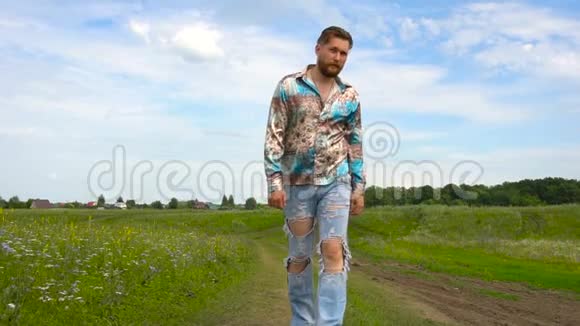 夏天一个留着胡子穿着破牛仔裤的男人在田野里散步视频的预览图