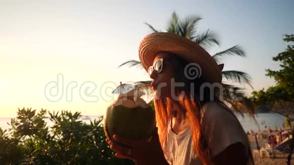 迷人的年轻混合种族旅游女孩喝新鲜泰国椰子水可可在海滩与美丽的日落4K视频的预览图