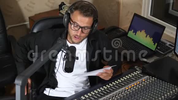 男性电台主持人带着一张纸对着眼镜对着收音机演播室设备附近的麦克风讲话视频的预览图