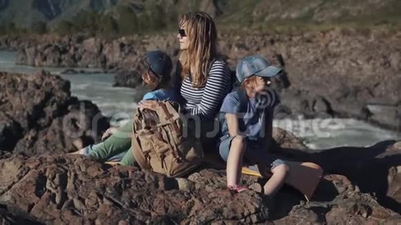一个年轻的旅行者妈妈和她的孩子坐在靠近山河的悬崖边游客的家人视频的预览图