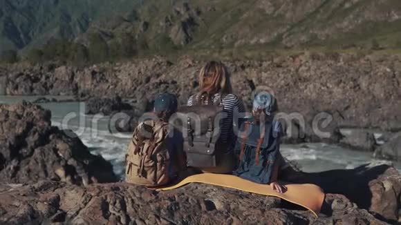 一个年轻的旅行者妈妈和她的孩子坐在靠近山河的悬崖边游客的家人背面视图视频的预览图