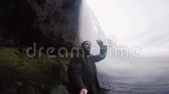 年轻的快乐男子与自拍棒拍摄在戈普罗相机强大的瀑布Seljalandsfoss在冰岛雾天视频的预览图