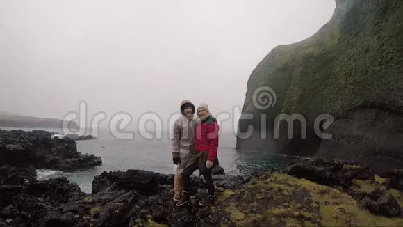 年轻的旅行夫妇穿着雨衣站在海边享受这次旅行游客男人和女人视频的预览图